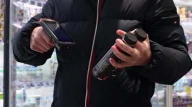 Genç bir adam, kapalı bir dükkanda dikilirken el tarayıcısı ile plastik şişe yoğurt içerken barkodu tarıyor. Çevrimdışı alışveriş konsepti..