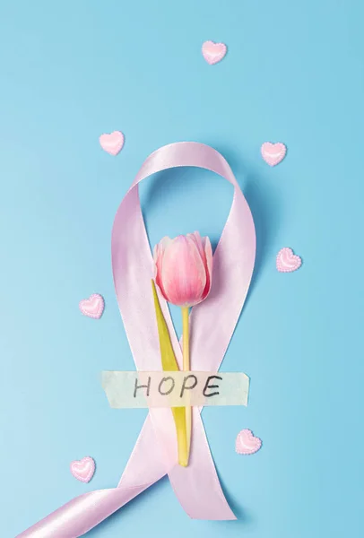 One pink ribbon emblem with a tulip attached with sticky tape with the word hope and decorative hearts scattered nearby lie on a blue background, flat lay close-up. Concept for world cancer day, breast cancer.