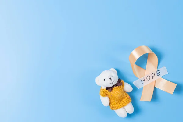 One yellow ribbon with sticky tape with hope and a toy bear lie on the right on a blue background with copy space on the left,flat lay close-up.Concept for World Childhood Cancer Day.