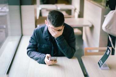 Koyu renk ceketli beyaz bir adamın portresi bir restoranın penceresinin yanındaki masada oturuyor ve eliyle yüzünü ovuyor, bir yandan cep telefonu tutuyor, yemek siparişini bekliyor, yakından yan görüş..