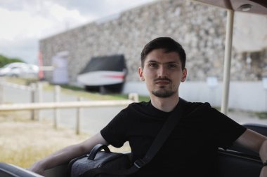 Yazın siyah tişörtlü, turist vagonunda oturan ve Etretat, Fransa 'da kameraya bakan beyaz tenli genç bir adamın yakın plan portresi..