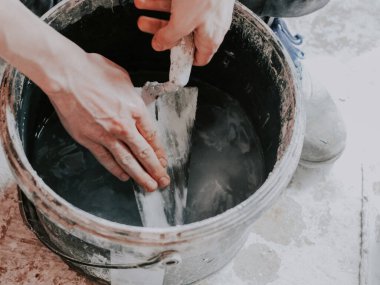 Beyaz bir gencin elleri siyah bir kovada kirli bir spatulayı yıkıyor. Dairenin zemininde temiz su var. Yakın çekimde. Bir spatulayı yıkamak, duvarları çürütmek,