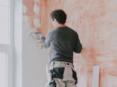 Gri tişörtlü, kollu, iş pantolonlu ve kıvırcık saçlı beyaz bir adam pencerenin yanındaki duvarda küçük bir spatulayla taze macunu uyguluyor.