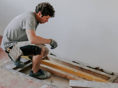 Kahverengi kıvırcık saçlı, beyaz üniformalı genç bir inşaat işçisi, yenilenmekte olan bir evin zemininde otururken şömine için yer ölçer.