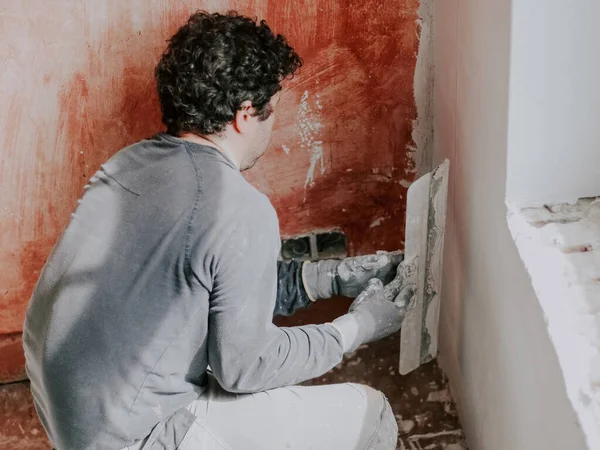 stock image A young caucasian guy in a gray t-shirt with sleeves and curly brown hair checks for evenness a fresh putty on the wall under the window holding a large spatula in his hands, close-up side view. The