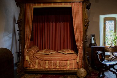 One antique bed with a red luxury canopy in a room in the medieval castle of Vianden in Luxembourg city, side view, close-up with a blurred background. Medieval lifestyle concept.