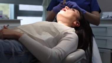 Mavi üniformalı ve eldiven takmış bir dişçi aletleri alır ve bir sandalyede, yan görüntüde yatan genç ve güzel bir kızın dişetlerini ve dişlerini temizlemeye başlar..