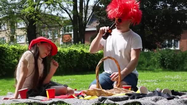 一名戴着比利时国旗太阳镜 头戴红色假发 头戴啤酒的年轻迷人男子和小女儿坐在公园的草坪上庆祝比利时独立日 比利时日概念 — 图库视频影像