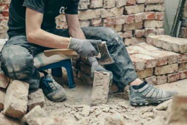 Siyah üniformalı, tanınmayan genç beyaz bir adam, baltayla kirli bir çimento kalıntısını temizliyor, bacaklarının açık olduğu küçük bir bankta oturuyor ve tuğla yığını onarılıyor. Yan görünüm