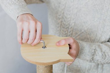 Hands of a young caucasian man in a knitted sweater manually twists a screw with a curly screwdriver into a board while assembling a cat scratching post against a white wall, close-up side view.Concept assembly of a scratching post, cat furniture.