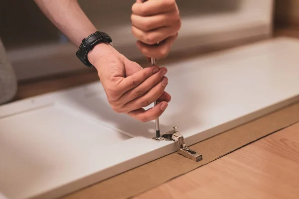 stock image Young caucasian unrecognizable guy hand tightens a screw with a screwdriver on a white wooden wardrobe door, sitting on the floor, close-up view from below with selective focus. Furniture assembly concept.