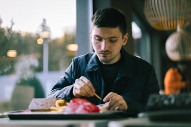 Koyu renk ceketli beyaz tenli bir gencin portresi bir lokantada bir masada oturur ve fast food yer, sosa patates batırır, yakın plan çekimler yapar..