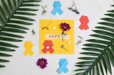 Çok renkli kağıt kurdeleler, leylak gerbera çiçekleri, umut kelimesinin geçtiği yapışkan bant, kenarları etrafında haplar ve palmiye dalları beyaz taştan bir arka planda, düz bir şekilde uzanıyor. Dünya Kanser Günü konsepti, bitkisel tıp.