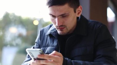 Koyu renk ceketli, beyaz tenli genç bir adam bir lokantanın penceresinin yanındaki masada oturur ve dikkatle telefon ekranına bakar. Parmaklarını ekrandan geçirir..