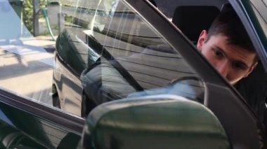 Koyu renk ceketli genç beyaz bir adam yolcu tarafındaki kapının camını mavi bir bez parçasıyla yıkıyor ve güneşli bir bahar gününde umumi oto yıkama yerinde kapalı tutuyor..