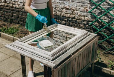 Yeşil şortlu ve eldivenli, tanınmayan genç bir beyaz kız bir ayna çerçevesini geniş bir fırçayla boyuyor. Bir yaz günü evin arka bahçesinde dikilirken yansıtıyor..