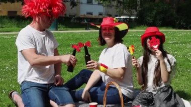 Belçika bayrağı takmış, kızlarıyla birlikte bira içen Belçika Bağımsızlık Günü 'nü kutlamak için parkta çimlerde oturmuş eğlenen ve gülen genç bir çift. Belçika Günü konsepti.