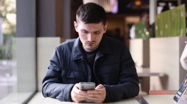 Koyu renk ceketli, beyaz tenli genç bir adam bir lokantanın penceresinin yanındaki masada oturur ve dikkatlice ekrandaki telefonun ekranına bakar. Ekranda parmağını geçirir. Yemek siparişini bekler. 