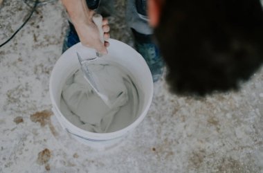 Beyaz tenli genç erkek, beyaz ıslak macunu küçük bir kovada inşaat spatulasıyla karıştırırken kapalı alanda kirli zemin üzerinde duruyor, üst manzara seçici bir odak ile kapanıyor. Ev yenileme konsepti, inşaat işi.
