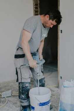 Beyaz macunu beyaz bir kovayla karıştıran beyaz tenli genç, beyaz bir erkek, kirli bir yerde, kapalı bir odada duruyor, üst tarafı kapalı, seçici bir odak noktası var. Ev yenileme konsepti, inşaat işi.