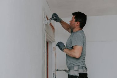 Gri üniformalı ve eldivenli genç bir beyaz tenli esmer, inşaat ağını beyaz macunla kaplıyor, kapı girişinde bir merdiven üzerinde yan duruyor, aşağıdan seçici bir odak ile yakından bakıyor. Ev yenileme konsepti.