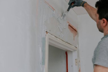 Gri üniformalı ve eldivenli genç bir beyaz esmer, inşaat ızgarasını spatula ile kaplıyor, kapı girişinde yarı açık duran bir merdiven, aşağıdan seçici bir odak ile yakın görüş. Ev kavramı