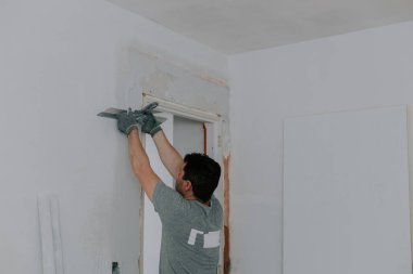 Gri üniformalı ve eldivenli genç bir beyaz esmer, inşaat ızgarasını spatula ile kaplıyor, kapı girişinde yarı açık duran bir merdiven, aşağıdan seçici bir odak ile yakın görüş. Ev kavramı