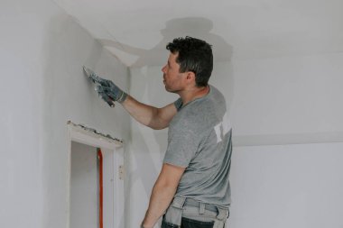 Gri üniformalı yakışıklı beyaz bir adam ve merdivenin üzerinde duran eldivenler akşamları duvarları spatula ile kaplıyor ve bir bina feneri açık ve yakın çekim mekanı kopyalıyor. İnşaat konsepti.