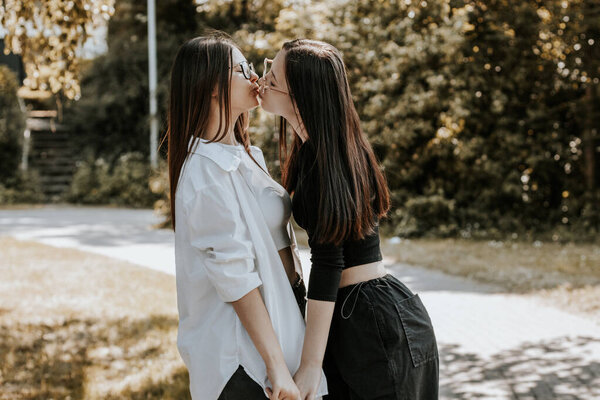 Portrait of two young beautiful Caucasian girls holding hands and kissing on the lips in a city park on a summer day, bottom view.