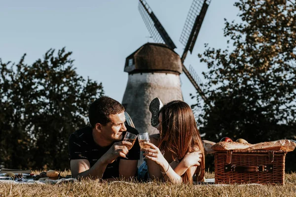 stock image One beautiful caucasian young couple lies on their stomachs on a bedspread with a wicker basket, fruits and tenderly look at each other, holding glasses of champagne in their hands in a park against the backdrop of an old windmill, close-up side view