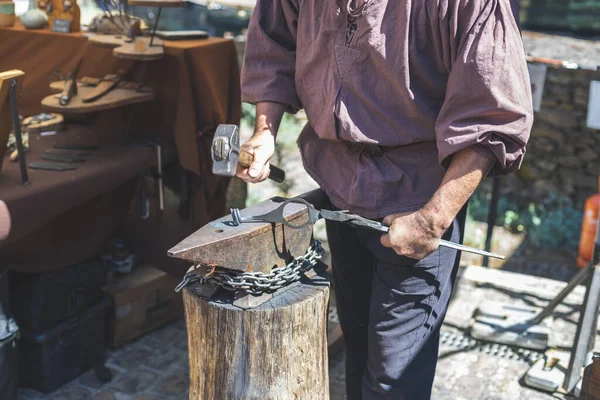 One middle-aged caucasian male blacksmith forges iron with a sledgehammer, standing behind a wooden stump on the street of Luxembourg city medieval castle, side view, close-up. The concept of medieval needlework, artisanal.
