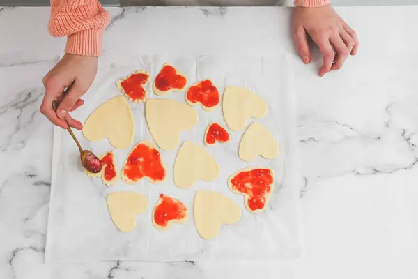 The hands of a young caucasian baker girl are smeared with strawberry jam, hearts of different shapes and sizes are cut out of raw dough on a white parchment at a marble table, flat lay close-up. Cooking at home, homemade cookies and valentine's day