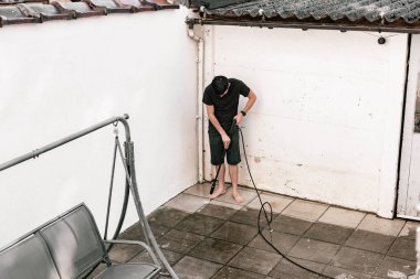 Siyah elbiseli genç beyaz bir adam evinin arka bahçesinde bir fayans zeminini bir kerpetenle yıkıyor..