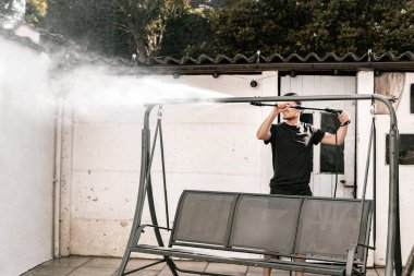 Siyah elbiseli esmer beyaz bir adamın portresi çıplak ayakla evinin arka bahçesinde çıplak ayakla salıncakta salıncak yıkarken basınçlı su altında bir jet ile yan tarafından yakın çekim görüntüsü olan bir kercher..