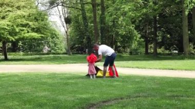 Kırmızı peruklu ve babasıyla birlikte giyinmiş küçük bir kız, güneşli bir yaz gününde parkın çimlerinde Belçika bayrağını sallıyor ve koşuyor. Belçika Günü konsepti.