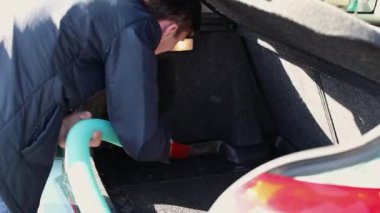 Beyaz tenli genç bir adam güneşli bir bahar gününde açık bir araba yıkamada arabasının bagajını süpürüyor..