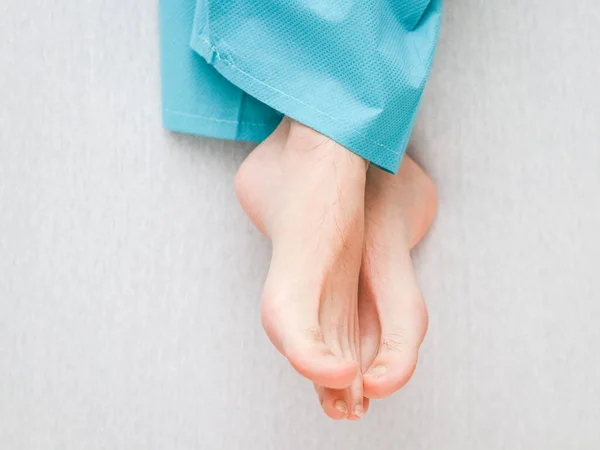 Clean and crossed legs of a young Caucasian patient in disposable pajamas lying on a bed with white linens and a blurred background, close-up top view. Body part concept.