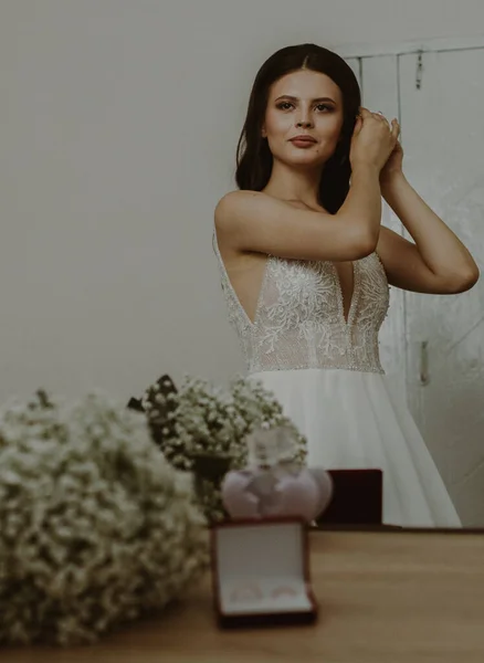 One beautiful caucasian brunette bride dresses up in front of a mirror while adjusting her hairdo, close-up side view.