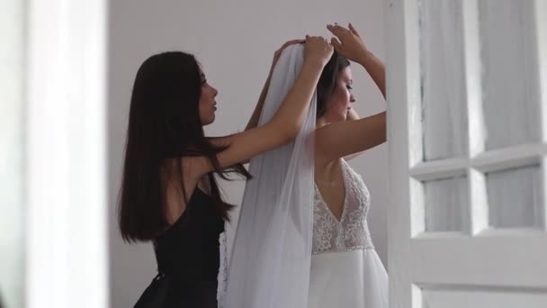 美しいブルネットの花嫁は花嫁が鏡の前で部屋に立っているベールの上に置くのを助けます 側面の眺めのクローズアップ ハンドヘルドショット — ストック動画
