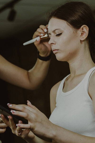 Portrait of a young beautiful Caucasian girl sitting on a chair in a beauty salon with a cell phone in her hands, the master is applying concealer with a brush on her face, side view.