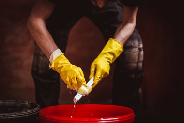 Siyah elbiseli, sarı lastik eldivenli, beyaz, tanınmayan genç bir adam. Bir kova sabunlu su ve süngerle pencere çerçevelerini yıkıyor. Tadilatın devam ettiği bir odada duruyor.,