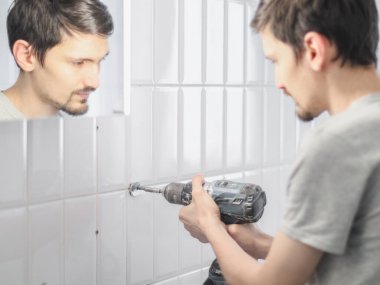 Genç, yakışıklı, beyaz tenli bir adam banyodaki eski metal bir havlu askısının vidalarını söker. Elinde matkapla aynaya yansıtır..