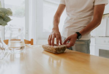 Tanınmayan genç bir beyaz adam buğday tohumlarını iki eliyle bir kabın içine koyup ahşap bir masada duruyor..