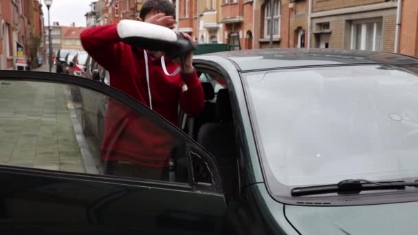 赤いフーディーのハンサムな若い白人男性は 街路の暗い緑色の車に白い後部鏡を設置し サイドビューのクローズアップを遅い動きで — ストック動画