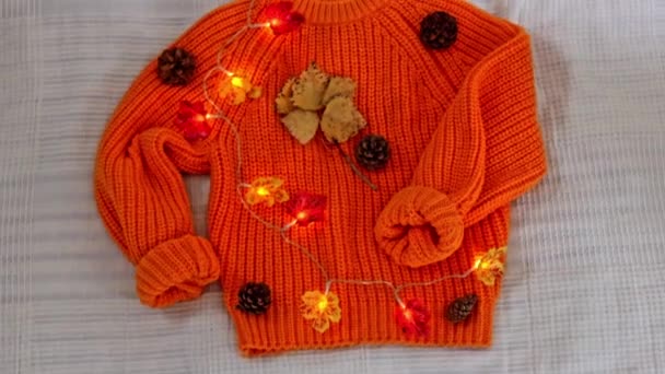 ピネコーンと秋のポプラの葉の燃えるガーランドが付いている1つのオレンジ編まれたセーターは 白いベッドスプレッド 平らな層のハンドヘルドの遅い動きのクローズアップが付いているベッドの上にあります — ストック動画
