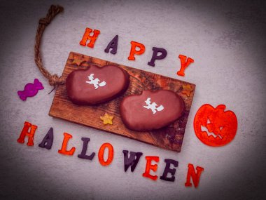 Balkabağı şeklindeki çikolatalı kurabiyeler, üzerinde renkli jöle şekerlerin yazılı olduğu tahta bir tahtanın üzerinde uzanıyorlar. Gri arka planda mutlu cadılar bayramı kutlaması, düzgünce uzanın. Cadılar Bayramı gıda kavramı..