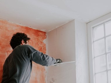 Üniformalı ve inşaat eldivenli beyaz saçlı genç bir adam pencerenin kenarına bir duvar yapıştırıyor, bir merdiven üzerinde duruyor, alt tarafı yakın plan. Daire yenileme konsepti.