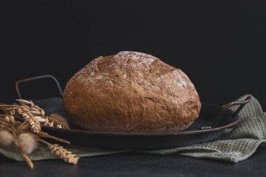 One round rye bread in a vintage metal plate with a kitchen napkin and ears lie on a black stone background, close-up side view.Bread baking concept.