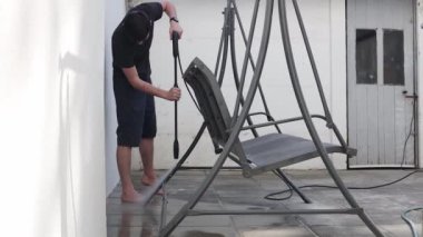 Genç, esmer, beyaz bir adam bir yaz akşamı evinin arka bahçesinde bir Karcher ile salıncağını yıkıyor..