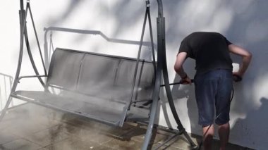 Koyu renk elbiseli esmer, beyaz bir adam sırtüstü duruyor ve bahçedeki salıncağın koltuklarını güçlü bir su basıncıyla yıkıyor. Bir yaz akşamı evinin küçük arka bahçesinde bir Karcher kullanıyor..